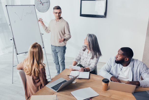 multicultural colleagues sitting at table and looking at businessman near flipchart with goals lettering  - Photo, Image